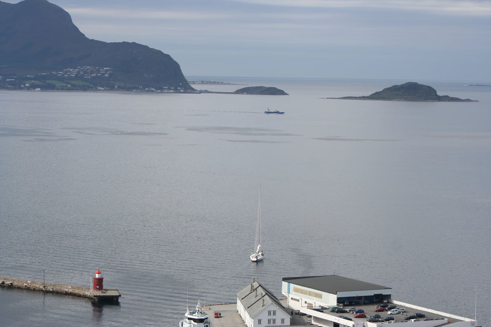 Grå, stille sjø utenfor Ålesund med Godøya og Havstein, en båt i fart og et lite fyr.