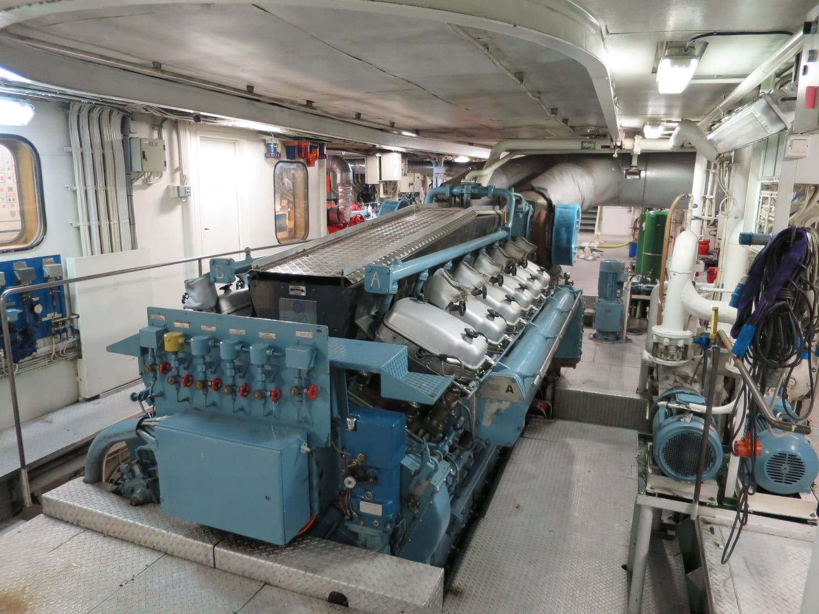 Et ryddig maskinrom med en stor blå dieselmotor, rørføringer og pumper under lysrør.