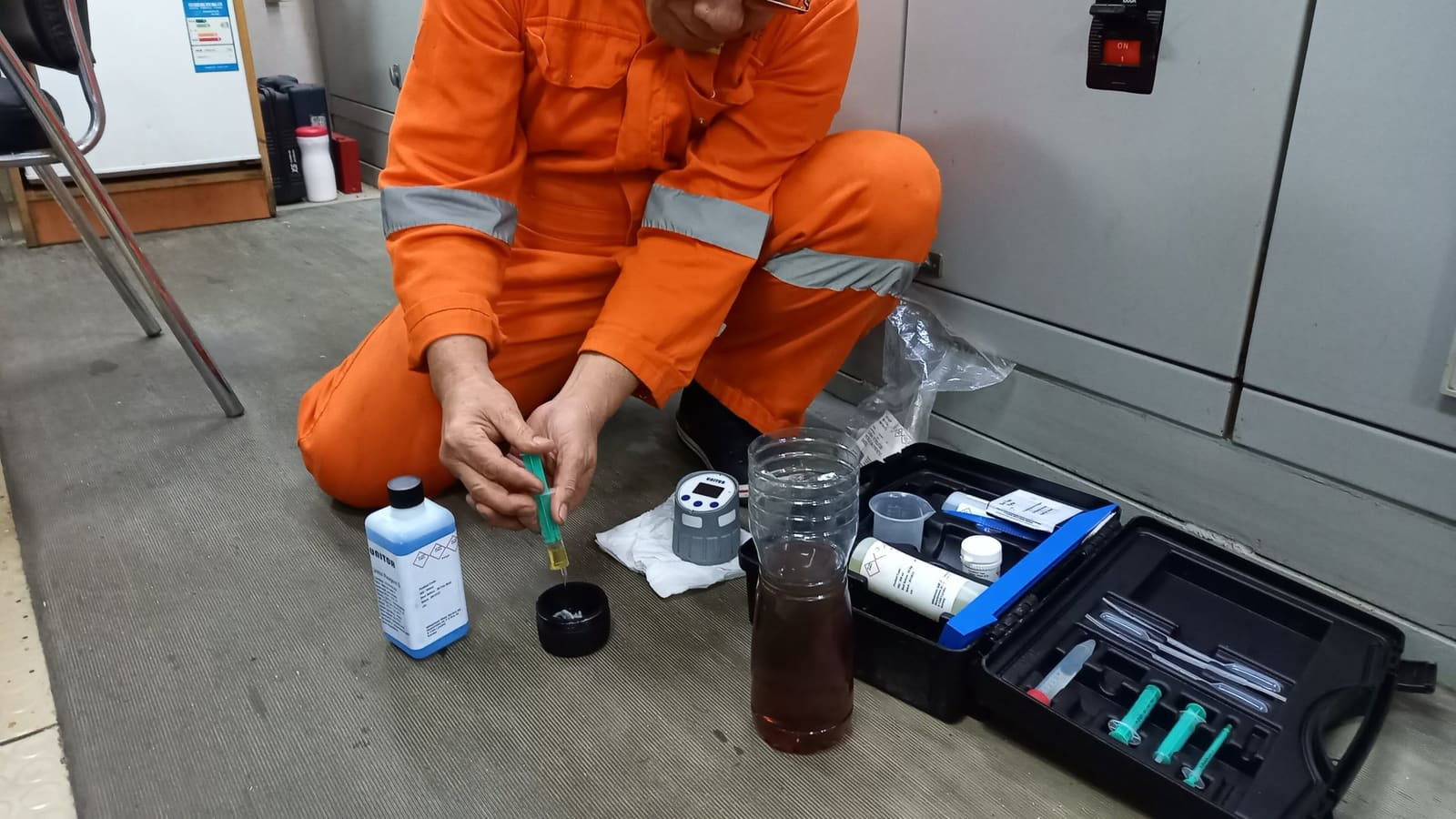 Et besetningsmedlem i oransje kjeledress som kneler på dekk og tester en oljeprøve med et testsett.