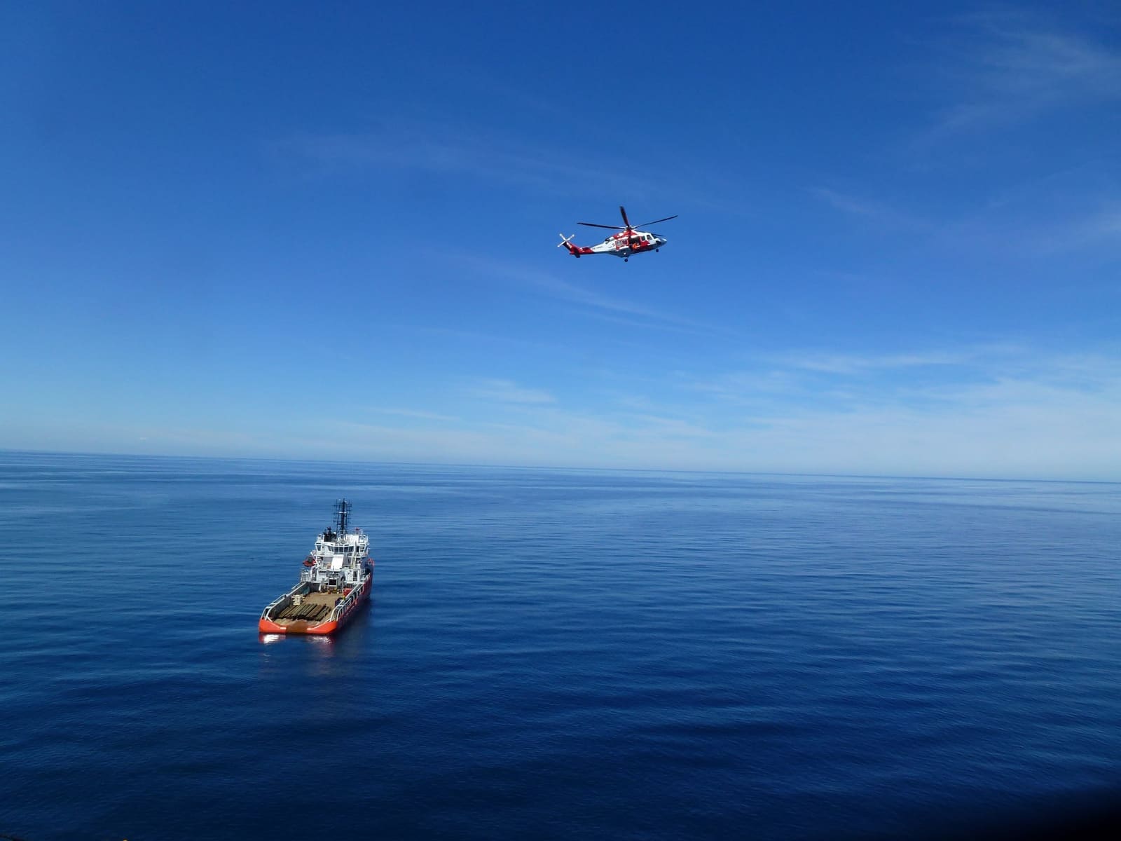 Et offshorefartøy på dypblått, åpent hav sett fra en rigg, med et helikopter som passerer over.