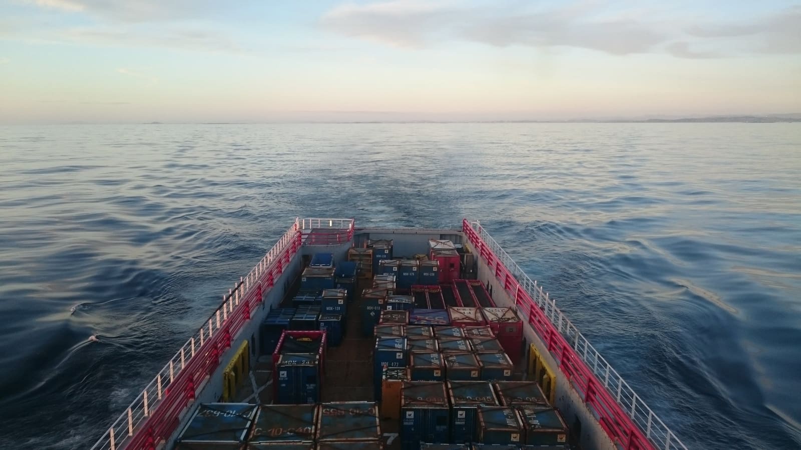 Lastedekket på et plattformforsyningsskip, lastet med containere, på vei ut over rolig sjø i skumringen.